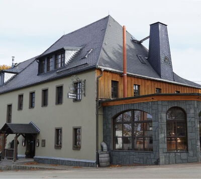 Traditionelles Gebäude mit Schieferdach, Holzfassade und steinernem Anbau mit großen Rundbogenfenstern.