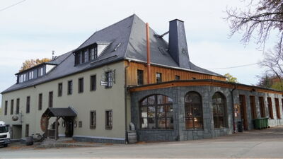 Traditionelles Gebäude mit Schieferdach, Holzfassade und steinernem Anbau mit großen Rundbogenfenstern.