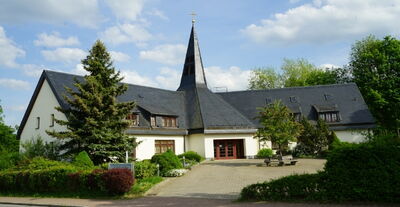 Kirche mit schiefergedecktem Spitzturm, Kreuz und gepflegtem Vorplatz mit Bepflanzung und Sitzbänken