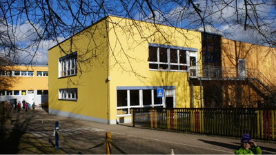 Moderner Zweckbau mit gelber Fassade, großen Fensterfronten und buntem Zaun im Eingangsbereich einer Schule
