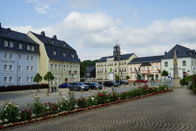 Historischer Marktplatz mit Rathaus, gepflasterter Fläche, Blumenbeeten und parkenden Autos vor pastellfarbenen Gebäuden.